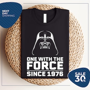 May include: Black t-shirt featuring a white graphic of Darth Vader's helmet and the text "ONE WITH THE FORCE SINCE 1976". The shirt has a crew neck and is made of a soft material. A perfect gift for Star Wars fans.