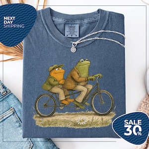 a blue t-shirt with a cartoon illustration of two frogs riding a bicycle, accompanied by a price tag and a sale promotion.