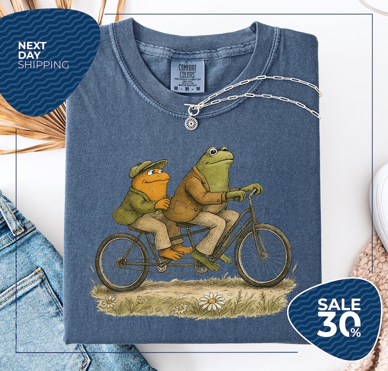 a blue t-shirt with a cartoon illustration of two frogs riding a bicycle, accompanied by a price tag and a sale promotion.