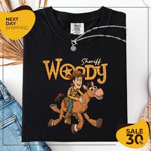 A black t-shirt with a cartoon image of Woody, a character from the Toy Story franchise, riding a brown horse. The t-shirt is displayed on a white background, and there are yellow and black sale signs indicating a 30% discount.