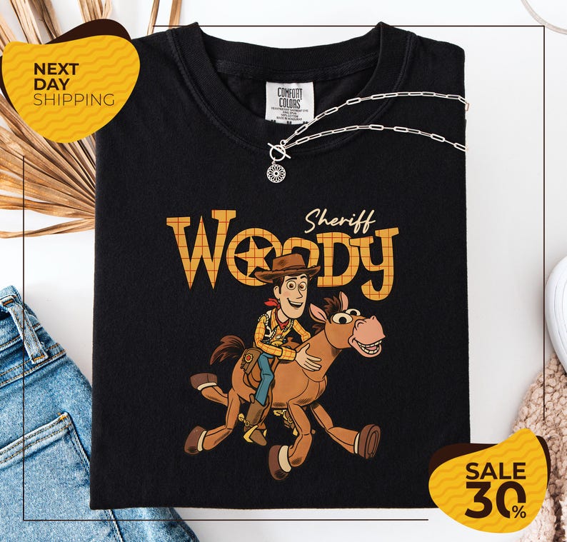 A black t-shirt with a cartoon image of Woody, a character from the Toy Story franchise, riding a brown horse. The t-shirt is displayed on a white background, and there are yellow and black sale signs indicating a 30% discount.