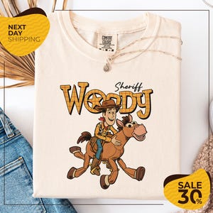 a white t-shirt with a cartoon image of Woody, a character from the Toy Story movie, riding a brown horse. The t-shirt is displayed on a white background, and there are yellow and black sale signs indicating a 30% discount.