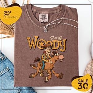 a brown t-shirt with a cartoon image of Woody, a character from the Toy Story movie, riding a horse. The t-shirt is displayed on a white background, and there are some jeans and a necklace visible in the background.