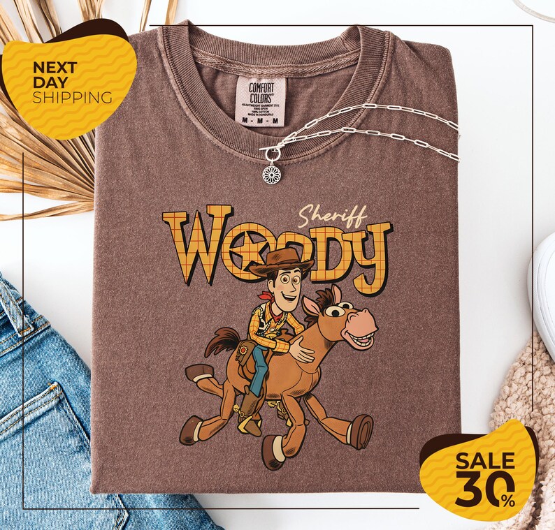 a brown t-shirt with a cartoon image of Woody, a character from the Toy Story movie, riding a horse. The t-shirt is displayed on a white background, and there are some jeans and a necklace visible in the background.