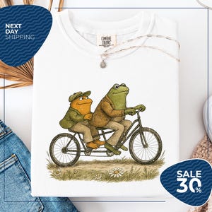 a white t-shirt with a drawing of two frogs riding a bicycle, accompanied by a sale sign indicating a 30% discount.