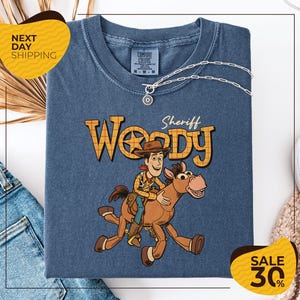 A blue t-shirt with a cartoon image of Woody, a character from the Toy Story franchise, and a sheriff badge on the front. The t-shirt is displayed on a white background, with a yellow sign indicating "Next Day Shipping" and a sale tag indicating a 30% discount.