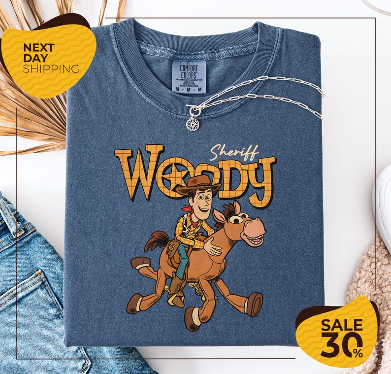 A blue t-shirt with a cartoon image of Woody, a character from the Toy Story franchise, and a sheriff badge on the front. The t-shirt is displayed on a white background, with a yellow sign indicating "Next Day Shipping" and a sale tag indicating a 30% discount.