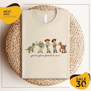 a beige t-shirt with a graphic design of five cartoon characters from the Toy Story franchise, including Woody, Buzz Lightyear, Timmy, and a horse. The text below the characters reads "you've got a friend in me!"
