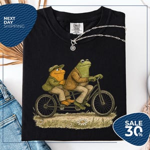 a black t-shirt with a whimsical illustration of two frog characters riding a bicycle, accompanied by a necklace and a tag. The t-shirt is displayed on a white background, and there is a sale sign indicating a 30% discount.