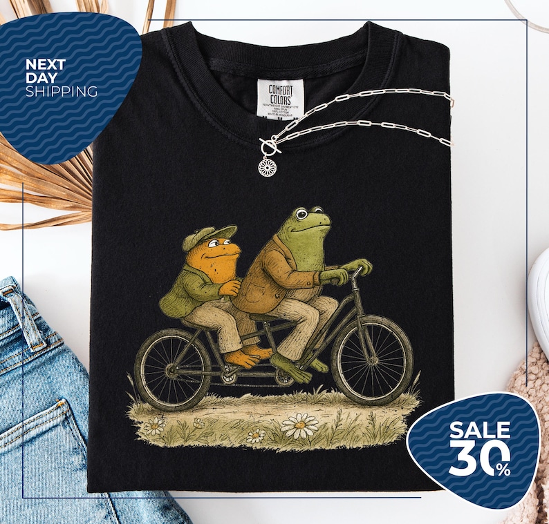 a black t-shirt with a whimsical illustration of two frog characters riding a bicycle, accompanied by a necklace and a tag. The t-shirt is displayed on a white background, and there is a sale sign indicating a 30% discount.