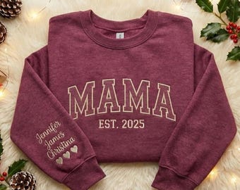 May include: A maroon sweatshirt with the word "MAMA" in white collegiate lettering and "EST. 2025" below. The left sleeve has embroidered names and hearts. The sweatshirt is on a fluffy white surface with holiday decorations.