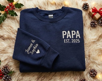 May include: Navy blue sweatshirt with the word "PAPA" and "EST. 2025" embroidered in white. The left sleeve has the names "James & Sophia" and a heart. The sweatshirt is on a faux fur surface with holiday decorations.