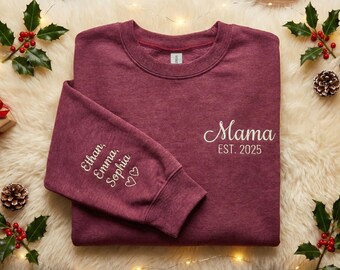 May include: A maroon sweatshirt with the word "Mama" and "EST. 2025" embroidered in white. The left sleeve has the names "Ethan, Emma, Sophia" and two hearts embroidered in white. The sweatshirt is on a fluffy white surface with holiday decorations.