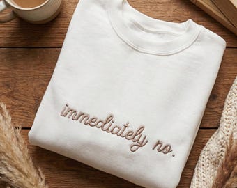 May include: Cream-colored sweatshirt with the embroidered phrase "immediately no." The crew neck sweatshirt is folded on a wooden surface, with a mug of coffee, a book, and decorative elements in the background.