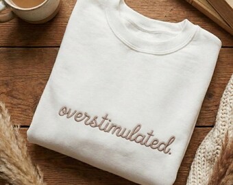 May include: Cream-colored sweatshirt with the word "overstimulated" embroidered in a cursive font. The sweatshirt is folded on a wooden surface, with a mug of coffee, a book, and decorative elements.