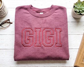 May include: A maroon sweatshirt with the name "GIGI" embroidered in red block letters. The sweatshirt is folded neatly on a white wooden surface, with a coffee mug, glasses, a succulent, and a framed photo nearby.