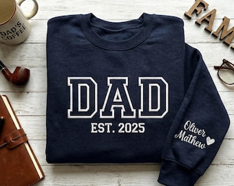 May include: Navy blue sweatshirt with the word "DAD" in white block letters and "EST. 2025" below. The sleeve has the names "Oliver Mathew" and a heart. A coffee mug, pipe, journal, and glasses are also visible.