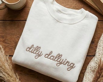 May include: Cream-colored sweatshirt with the embroidered phrase "dilly dallying" in a cursive font. The sweatshirt is folded on a wooden surface, with a mug of coffee and a book in the background.