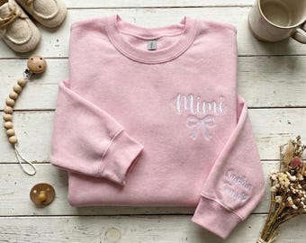 May include: A light pink sweatshirt with the word "Mimi" embroidered in white script, along with a bow design. The sleeve has the name "Sophia" embroidered. The image also includes baby shoes, a pacifier, and a wooden teether.