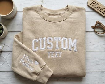 May include: A tan sweatshirt with white embroidered text reading "CUSTOM TEXT" across the chest. The left sleeve has embroidered text and a heart. A cup of coffee, glasses, and a succulent are also visible.