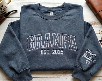 May include: A blue sweatshirt with the word "GRANPA" embroidered in white, along with "EST. 2025". The sleeve has the embroidered names "Oliver Mathew" and a heart. A mug, pipe, glasses, and a leather journal are also visible.