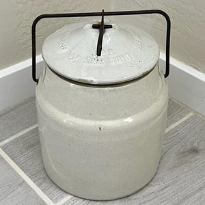 May include: A vintage-style, off-white ceramic jar with a lid and a dark metal handle. The lid has embossed text and a small metal cross-shaped handle. The jar is cylindrical with a slightly speckled finish, suggesting a rustic aesthetic.