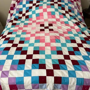 May include: A colorful handmade quilt with a checkerboard pattern. The quilt features squares of white, pink, teal, burgundy, and purple. The quilt is spread out on a bed, showcasing its design and craftsmanship.
