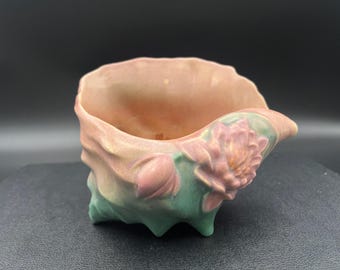 Może przedstawiać: Ceramiczna donica w odcieniach brzoskwini, różu i zieleni. Donica posiada rzeźbiony kwiatowy wzór z boku i falisty brzeg. Podstawa ma trzy małe nóżki. Ogólny kształt przypomina muszlę.