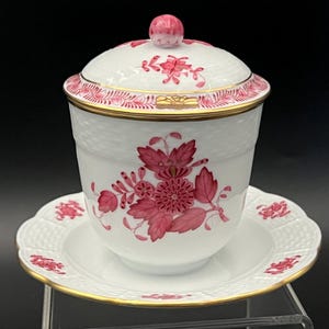 May include: A white porcelain teacup with a lid and saucer, adorned with a floral pattern in shades of pink and gold trim. The cup and saucer have a textured basketweave design. The lid features a pink knob.