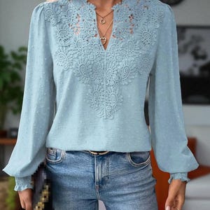 May include: A pale blue blouse featuring delicate lace embroidery on the neckline and front. The long sleeves are gathered at the cuffs and have a slight puff at the shoulders. The blouse is worn with blue jeans and a gold belt.