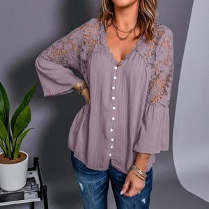 Op de afbeelding: Een mauve blouse met een V-hals en knoopsluiting. De mouwen zijn lang met uitlopende manchetten en de schouders en bovenmouwen hebben een bloemen kanten patroon. De blouse wordt gedragen met een blauwe spijkerbroek.