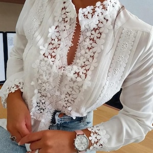 May include: A white blouse with long sleeves and detailed floral lace. The blouse has a high collar and lace trim on the cuffs and front. The design includes vertical pleats and a keyhole opening. A pink watch with a white face is visible.
