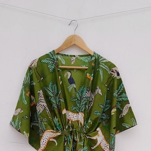 Peut inclure: Robe-kaftan verte avec un imprimé d'animaux de la jungle, dont des léopards, des zèbres et des toucans. La robe est suspendue à un cintre en bois. Le tissu est léger.
