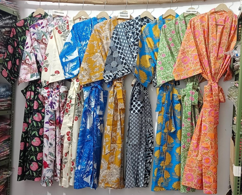 May include: A collection of colorful, patterned robes hanging on wooden hangers. The robes feature various floral and geometric designs in shades of blue, yellow, green, and orange. Some robes are tied with matching fabric belts. The robes are displayed against a white wall.
