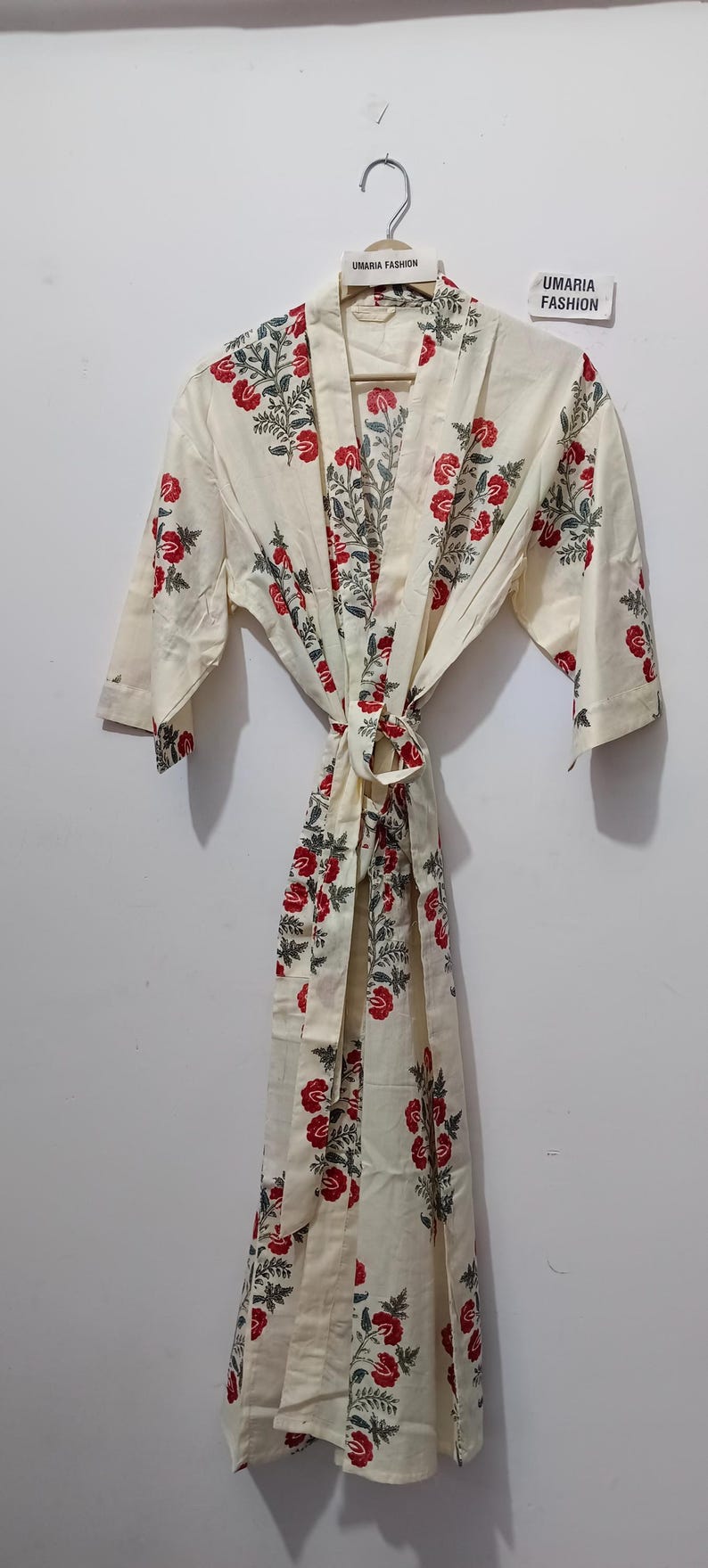 May include: Cream-colored floral print robe with red roses and green leaves. The robe has a tie closure and wide sleeves. The text "UMARIA FASHION" is visible on a tag. This robe is perfect for lounging.