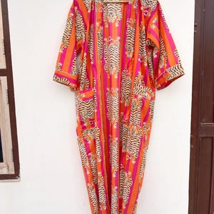 May include: A vibrant, open-front robe with a tiger stripe pattern in orange, pink, and black. The robe has three-quarter sleeves and two front pockets. It is hanging on a wooden hanger against a white wall.
