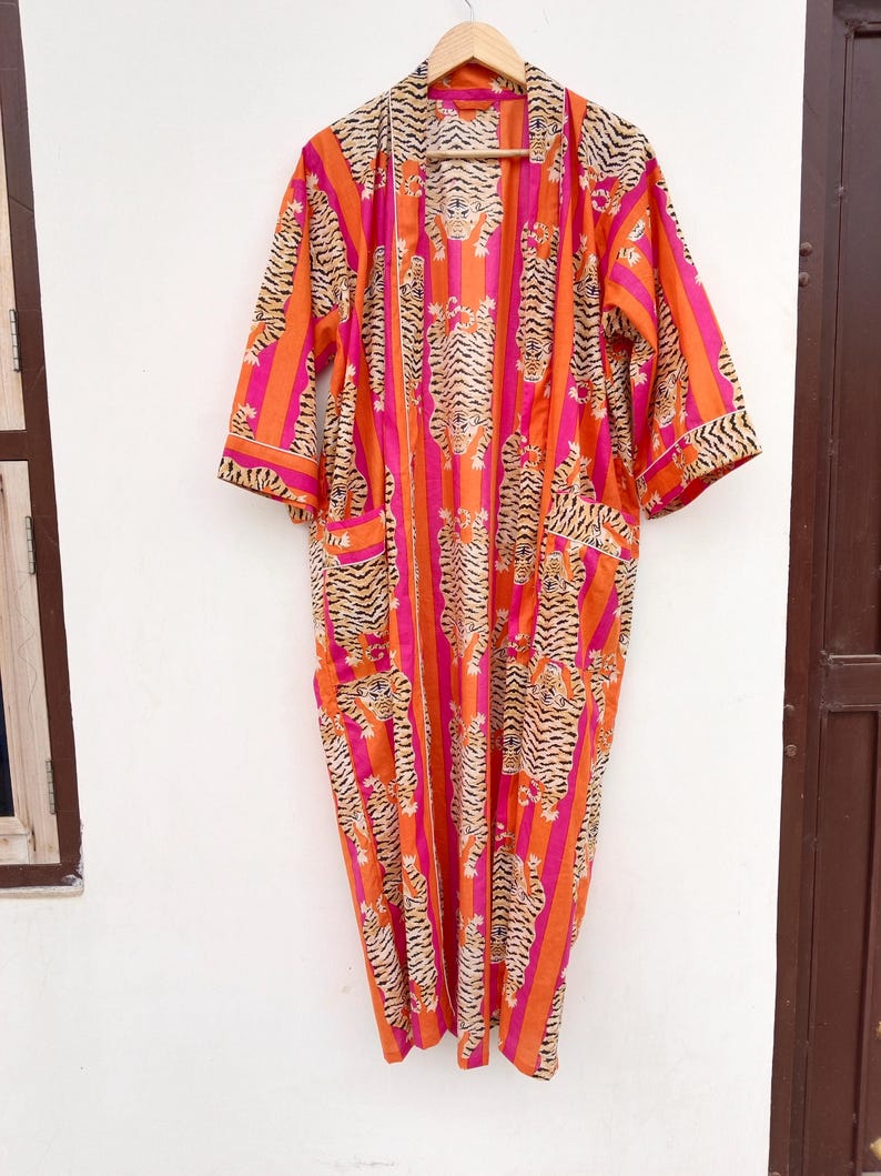 May include: A vibrant, open-front robe with a tiger stripe pattern in orange, pink, and black. The robe has three-quarter sleeves and two front pockets. It is hanging on a wooden hanger against a white wall.