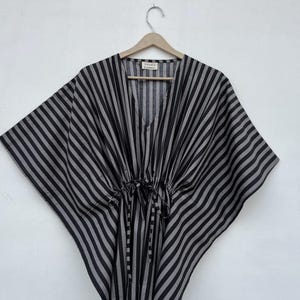 Könnte beinhalten: Ein schwarz-grau gestreifter Kaftan mit V-Ausschnitt, weiten Kimono-Ärmeln und einer Taille mit Kordelzug und Bindebändern. Das Kleidungsstück hat ein kleines weißes Etikett am inneren Halsausschnitt mit dem aufgedruckten Text "VILLAGE FASHION".