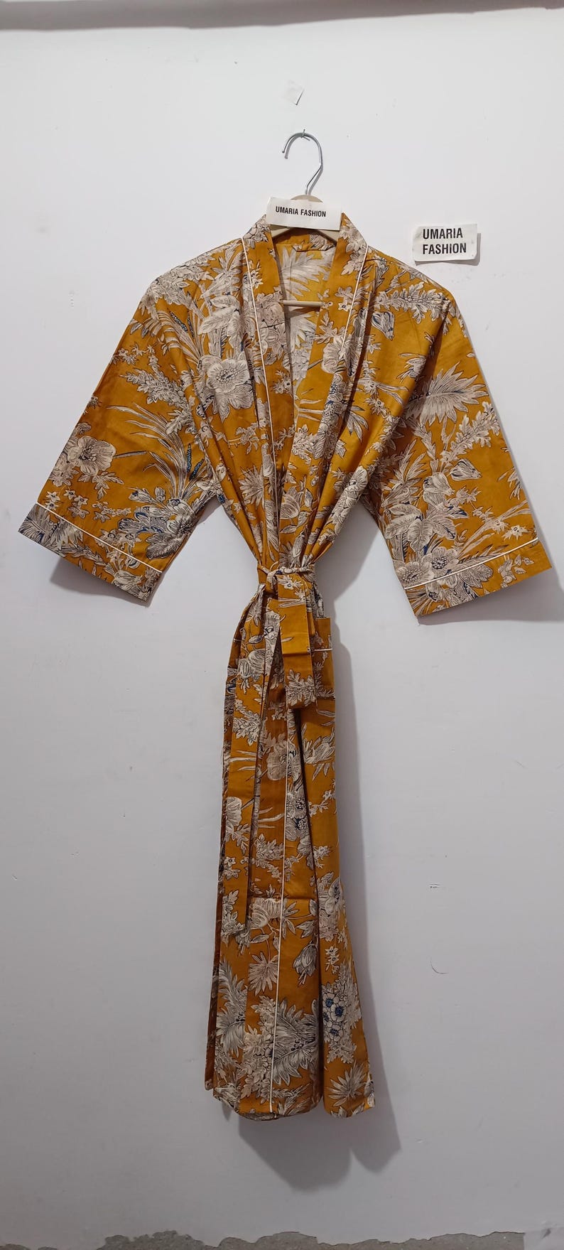 May include: Mustard yellow robe with a floral pattern in white and gray. The robe has wide sleeves, a tie belt, and white trim. The text "UMARIA FASHION" is visible on a tag and the hanger.