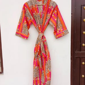 May include: A vibrant, patterned robe with a tiger stripe design in orange, pink, and tan. The robe is hanging on a wooden hanger against a white wall. The robe is tied at the waist with a matching belt.