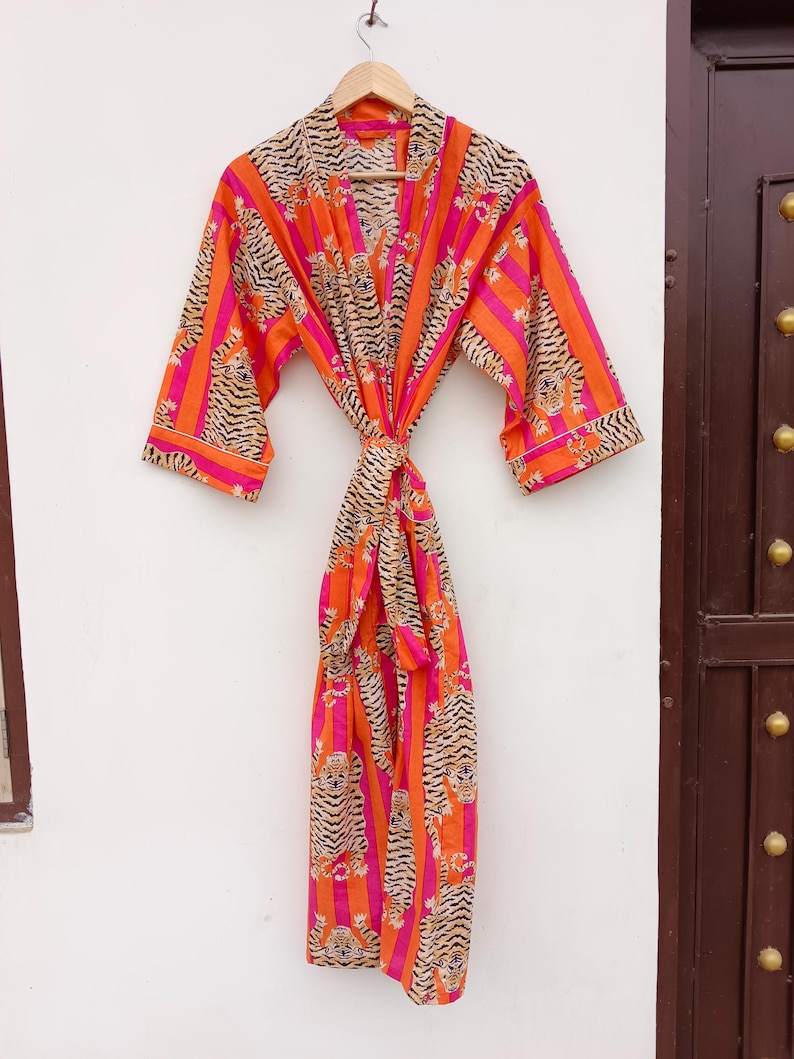 May include: A vibrant, patterned robe with a tiger stripe design in orange, pink, and tan. The robe is hanging on a wooden hanger against a white wall. The robe is tied at the waist with a matching belt.