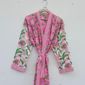 May include: A pink floral print dressing gown with a striped trim and tie. The gown features a pink and white floral pattern with green leaves. The sleeves and trim have a pink and white striped design. The gown is hanging on a wooden hanger.