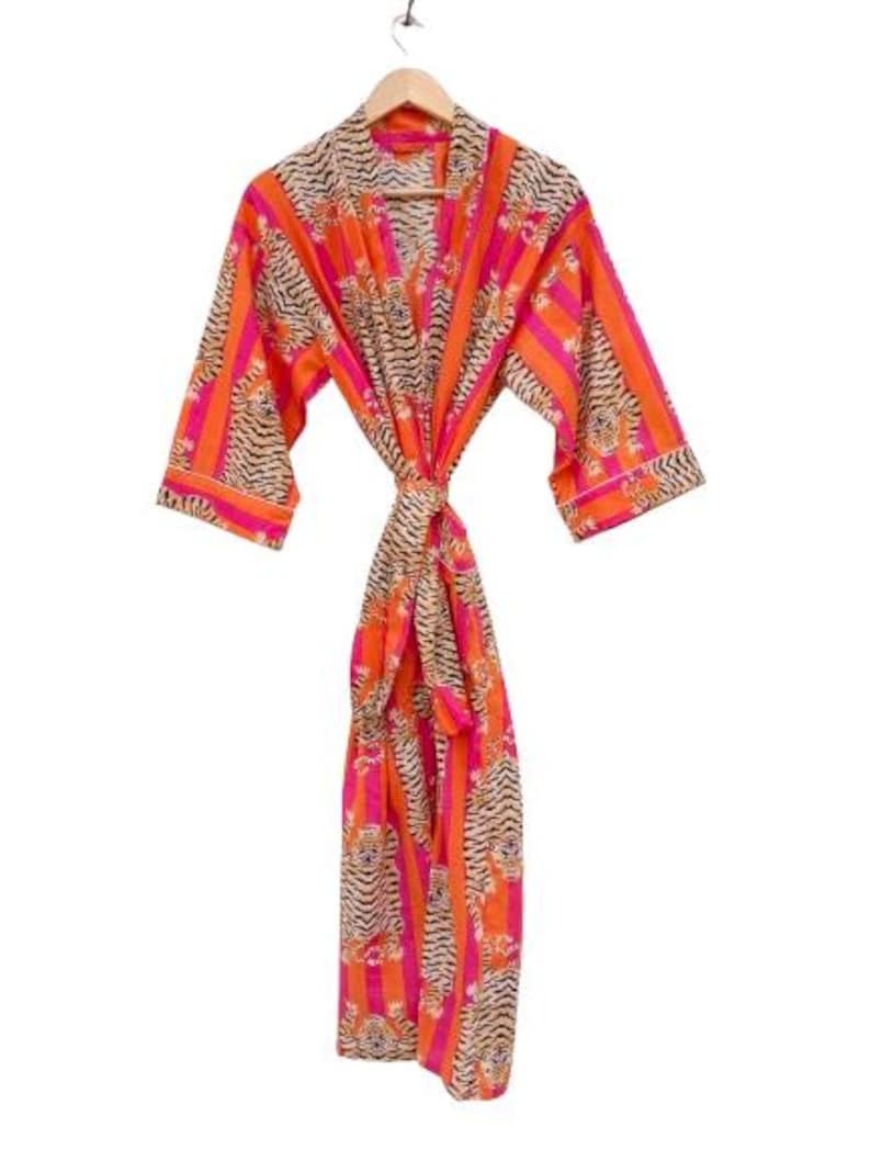 May include: A vibrant orange and pink striped robe with a tiger print design. The robe is hanging from a wooden hanger. The robe is tied at the waist with a matching belt. The robe is made of a lightweight fabric.