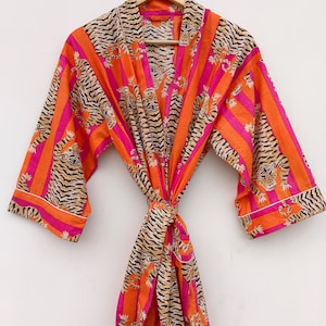 May include: An orange and pink patterned robe with a tiger stripe design. The robe is hanging on a wooden hanger. The robe has a tie closure and white trim.