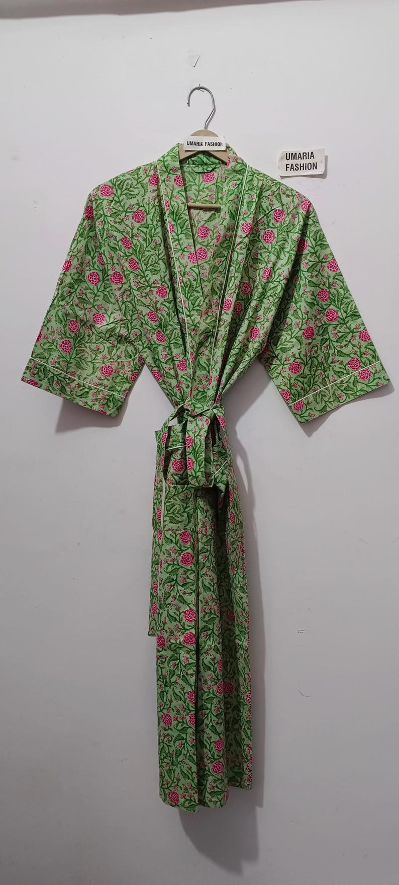 May include: A green floral print robe with pink flowers and white trim, hanging on a wooden hanger. The robe is tied at the waist. The brand name "UMARIA FASHION" is visible on the hanger and a tag.