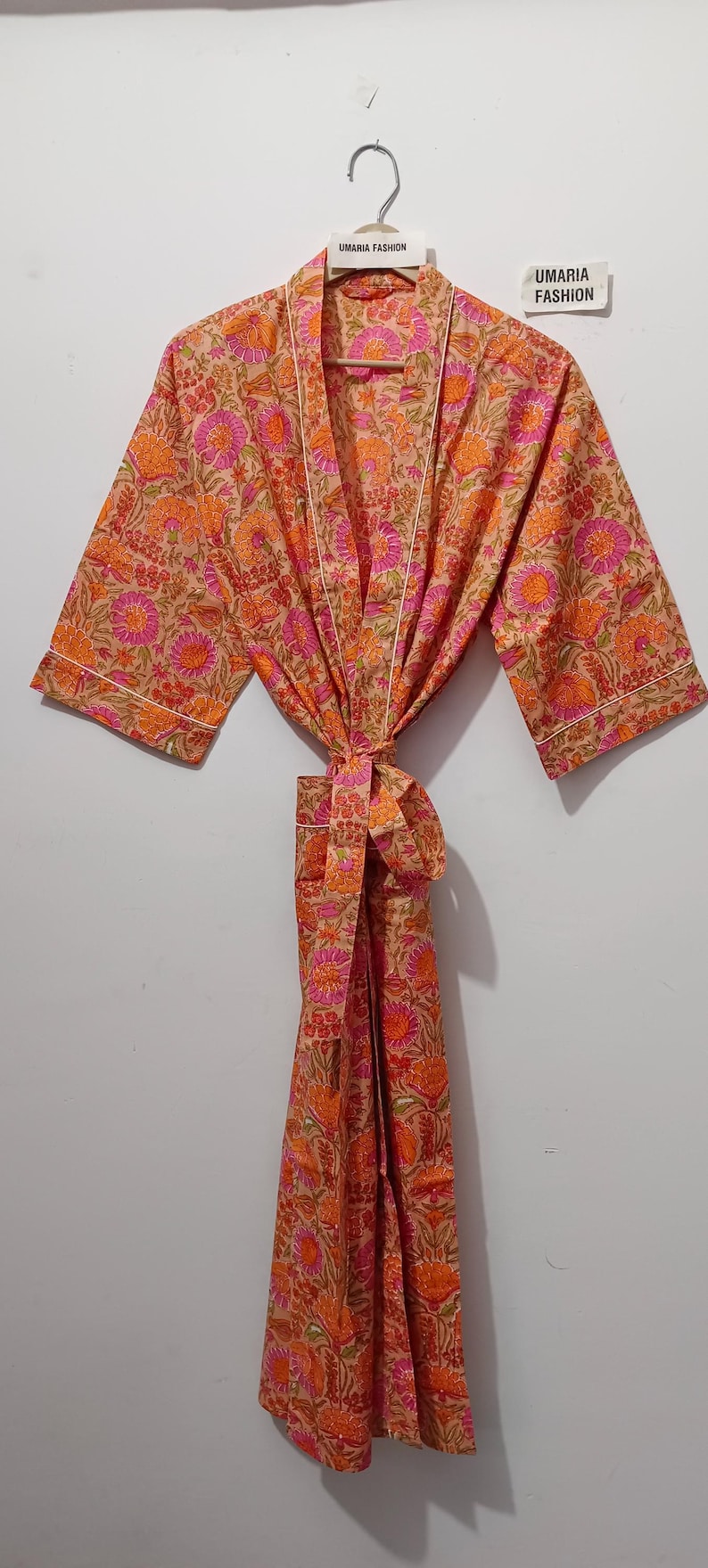 May include: A floral print robe in shades of orange, pink, and tan, hanging on a white wall. The robe features a tie closure and white trim. The brand name "UMARIA FASHION" is visible on the hanger.