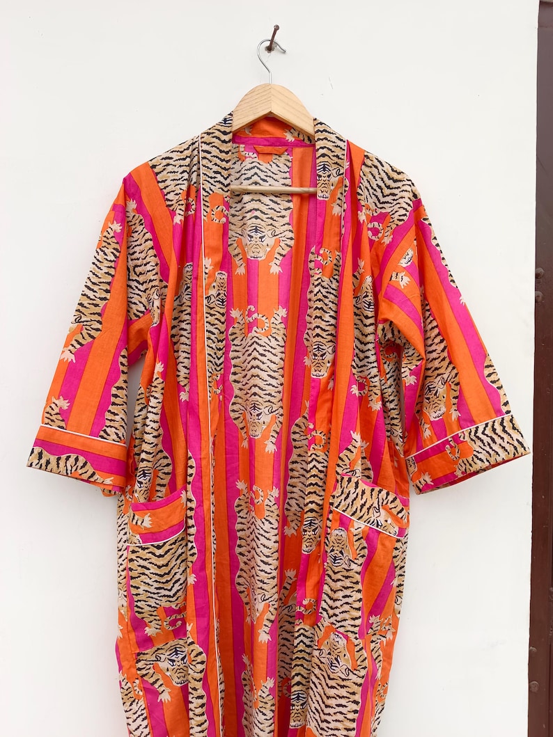 May include: An open-front robe with a vibrant tiger stripe pattern in orange, pink, and black. The robe features a striped design with tiger illustrations, pockets, and white trim. The robe is hanging on a wooden hanger.