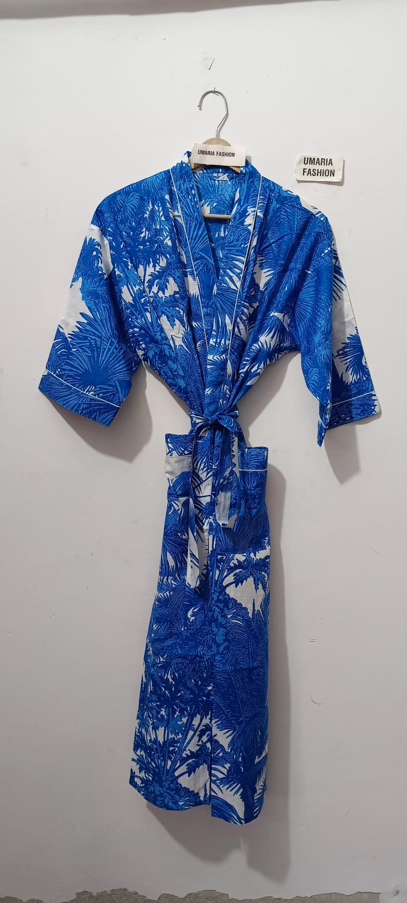 May include: Blue and white patterned robe with a palm tree design. The robe is hanging on a hanger and tied at the waist. The text "UMARIA FASHION" is visible on the hanger and a tag.