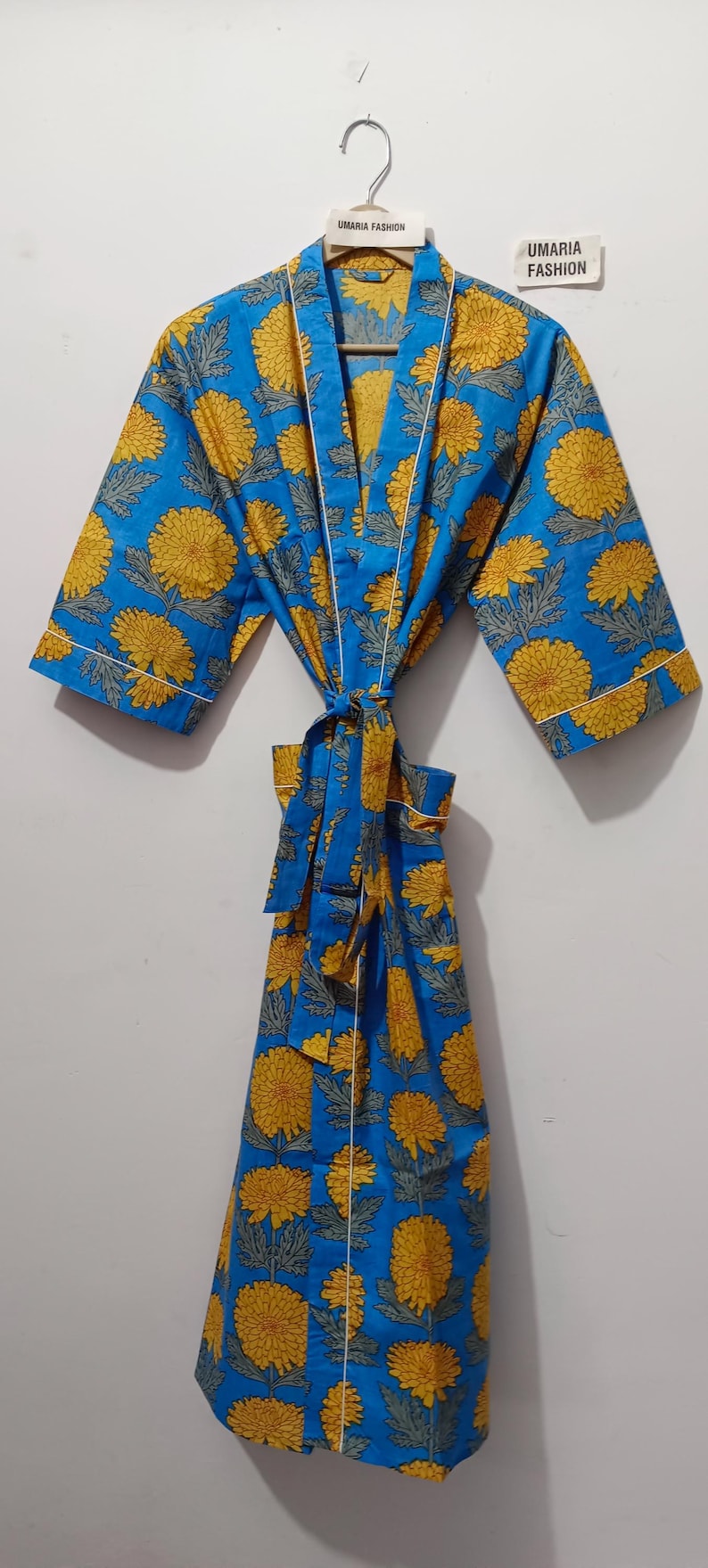 May include: A blue robe with a yellow floral pattern and white trim. The robe is tied at the waist with a matching belt. The text "UMARIA FASHION" is visible on a tag.