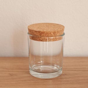 Op de afbeelding: Een heldere glazen pot met een kurken deksel. De cilindrische pot staat op een houten oppervlak. De ronde kurken deksel past goed bovenop. De achtergrond is een effen, lichtgekleurde muur.